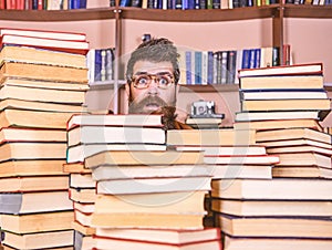 Man, nerd on surprised face between piles of books in library, bookshelves on background. Teacher or student with beard