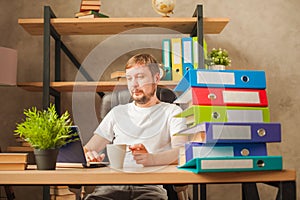 A man in the office behind a stack of folders and a laptop. Abused at work, making decisions at work. Male freelancer at home work