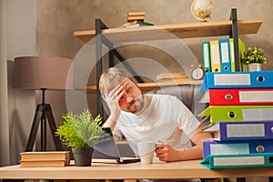 A man in the office behind a stack of folders and a laptop. Abused at work, making decisions at work. Male freelancer at home work