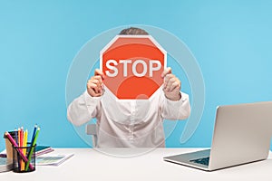 Man office worker in white shirt hiding face behind red stop traffic sign, sitting at workplace with laptop, symbol of prohibition