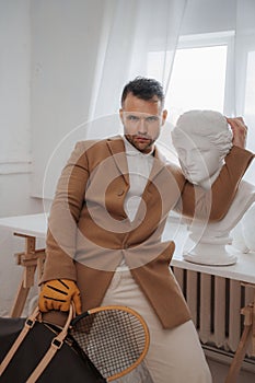 Man in old money style holding bag and tennis racket in sculptor's studio