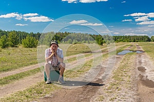 Man with old suitcase sitting on an country road and unsuccessfully waiting for any bus to pass by