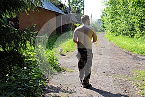 A man with an open torso is walking a small Pug dog on a hot summer day on a rural road.