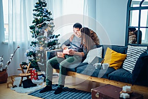 Man opening a gift box while her girlfriend sitting close to her on couch