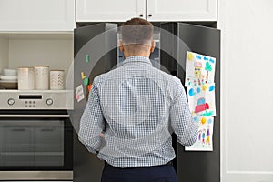 Man opening refrigerator door with child`s drawings, notes and magnets