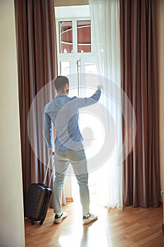 Man opens curtained window standing with suitcase in hotel room