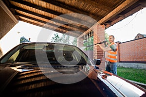 A man in orange vest washes his car with a large head of water from a karcher on open air. High pressure cleaning