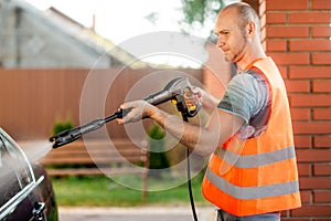 A man in orange vest washes his car with a large head of water from a karcher on open air. High pressure cleaning