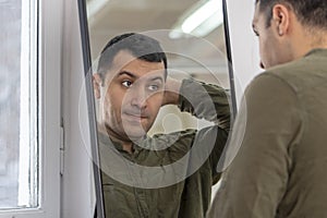 A man of oriental appearance 30-35 years old looks at his reflection in the mirror, scratches the back of his head, a light backgr