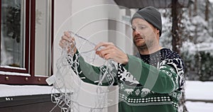 man outdoors in christmas sweater untangling string lights for house exterior decoration