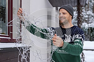 man outdoors in christmas sweater untangling string lights for house exterior decoration