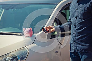 Man outdoors holding his new car keys and pink piggy money bank on the hood. Economic success, property insurance