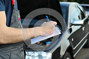 A man in overalls writes to a clipboard Mechanic. Car inspection