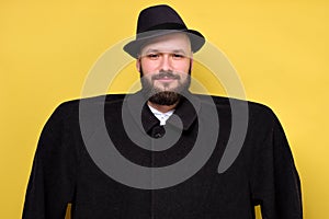 Man in oversize baggy black coat and hat smiling standing on yellow studio