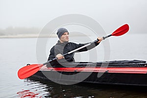 Man paddles red and black kayak with paddle in middle of river or lake in fall season. Autumn and winter kayaking, concentrated