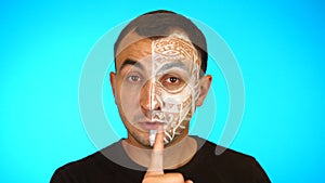 Man with painted face is asking for silence by putting finger on lips against blue background