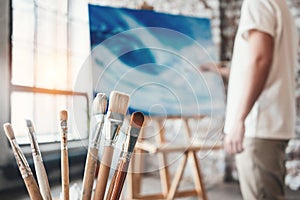 Man painter working in workshop with canvas on easel. Brushes close up on wooden table in studio. Flare effect