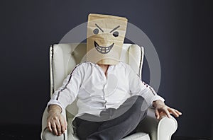 A man with a paper bag on his head, with a drawn angry smiley face, sits in a white chair.