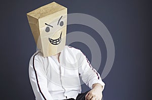 A man with a paper bag on his head, with a drawn angry smiley face, sits in a white chair.