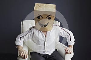 A man with a paper bag on his head, with a drawn angry smiley face, sits in a white chair.