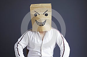 A man with a paper bag on his head, with a drawn angry smiley face, sits in a white chair.