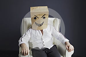 A man with a paper bag on his head, with a drawn angry smiley face, sits in a white chair.