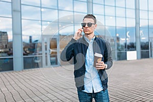 A man with a paper Cup of coffee, goes through the city, a handsome guy in stylish clothes and sunglasses, making a phone call