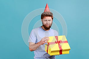 Man in party cone opening gift box and looking at camera with disappointed sad expression.