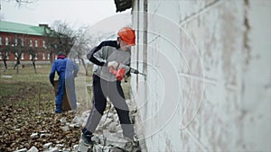 A man with a perforator breaking a wall outside. A man in a protective helmet. Demolition work