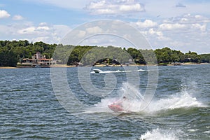 Man on Personal Water Craft landing in a spray of water after jumping a wake at the lake with speedboat and houses and boat docks