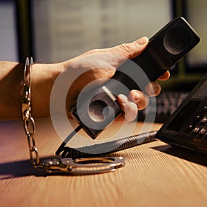 A man at the phone in handcuffs on his hands, close-up. Office desk in a dark night room with computer monitors