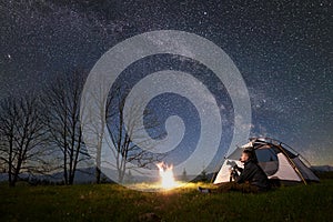Man with photo camera sitting alone near tourist tent at campfire under dark blue starry sky