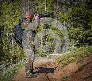 Man photographer in camouflage outfit with a backpack and tripod standing on a mountain forest trail and shooting