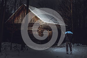 Man photographer standing in front of the abadoned and creepy cottage in forest in winter. Man on expedition is shinning with