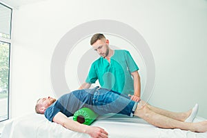 Man at the physiotherapy doing physical exercises with his therapist, they using a massage roll. A chiropractor treats patient`s
