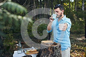 A man on a picnic in the woods checks the aroma of a drink in a glass.