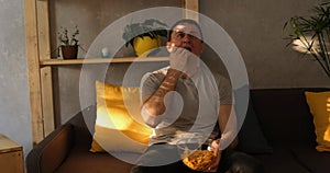 Man with a plate of crisps fits and sits down on the couch eating and watching TV. Natural evening light. Slow motion