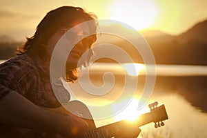 Man playing acoustic guitar outdoors with sunlight reflected on water surface at sunset lake