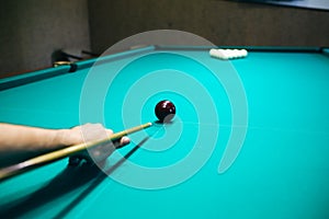 Man is playing billiard. Guy is holding pool cue in his hand. Small black cue ball is on the centre of the green table. Sports and