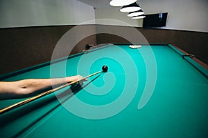 Man is playing billiard. Guy is holding pool cue in his hand. Small black cue ball is on the centre of the green table. Sports and