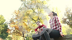 Man playing guitar while woman singing, sitting next to him on bench in park