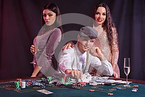 Man playing poker at casino sitting at table with stacks of chips, money, cards. Celebrating win with two women. Black