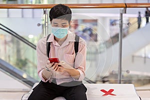 A man is playing a smartphone while sitting on a public shopping mall bench with caution social distancing signs. Selective focus