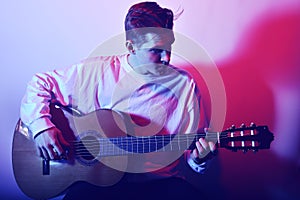 A man plays an acoustic guitar in neon red-blue light. A man learns to play the guitar, music, hobbies