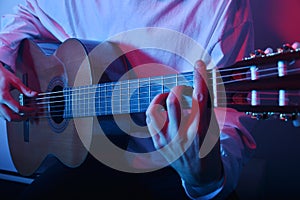 A man plays an acoustic guitar in neon red-blue light. A man learns to play the guitar, music, hobbies