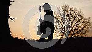 Man plays electric guitar and sings a lyric song in a field near the tree at sunset. silhouette