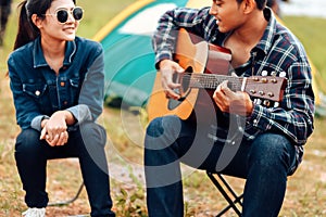 A man plays a guitar for a woman while camping beside the lake.