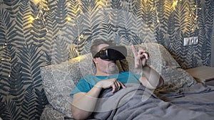 A man plays virtual games while lying in virtual reality glasses in the evening on a bed under a blanket against a