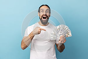 Man pointing at fan of dollars banknotes, looking at camera with big eyes and open mouth.