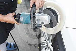 A man polishing the surface of a white sedan with an electric buffing machine. At an auto detailing shop